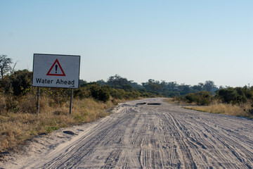 Water ahead warning sign alerts cars driving along an unpaved sandy road that the way has been flooded and it's interrupted. An hand-made arrow point to a detour through mopane trees. Botswana, Africa