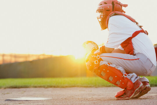 Baseball catcher squatting behind home plate backlit by sun.