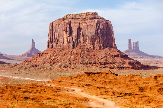 Picture of iconic Merrick Butte rock formation in Oljato-Monument Valley taken on a summer cloudy day from John Ford's point. The scenic drive dirt road with tourist cars winds through the red sand.