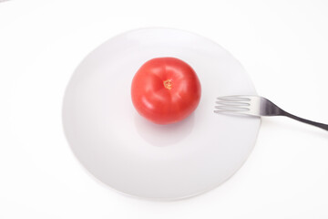 One single red ripe fresh raw tomato on white ceramic plate with a folk isolated on white, mediterranean diet concept