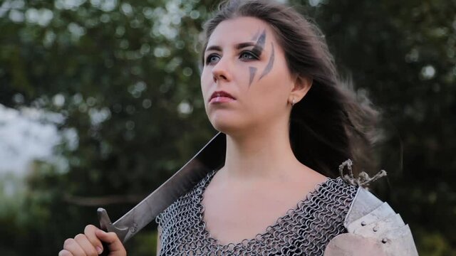 Portrait of a medieval woman warrior in chain mail armor with a plate shoulder pad with a shield and sword in her hands against the background of the forest bokeh. Wind blows the hair. Combat makeup.