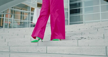Slow motion 4K fashionable green shoes and hot pink trousers. Stylish fashionista on heels walking down the marble stairs. Woman in pink outfit with white shopping bags with modern city on background