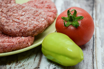 Ripe tomato and bell peppers on a background of raw homemade chopped beef burgers on a plate and a wooden table. BBQ preparation, beef products, traditional meat dish. Balanced diet. 