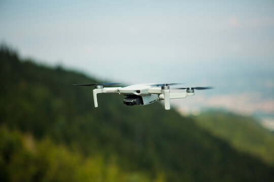 Mini white drone, in front of a hill in nature flying in a blue sky. The camera of the drone is facing towards focus. A flying drone captured from a high position.  A farest green backogrund with sky.