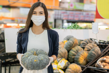 Beautiful woman wearing medical face mask and rubber glove holding pumpkin preparing for Halloween holiday at vegetable grocery department store. shopping at supermarket in new normal lifestyle