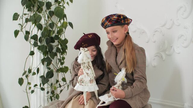 Adorable stylish girls in coats and berets posing with elegant vintage dolls. Cute stylish sisters sitting together and holding retro puppets in luxury interior