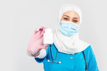 Fototapeta na wymiar friendly Muslim doctor or nurse in a hijab, mask, gloves offering a pill to the patient on a gray background