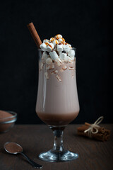 Hot cacao drink in tall glass with white marshmallow and cinnamon stick on dark brown wooden background with spoon at kitchen usually prepared at cold winter vacations. Vertical orientation
