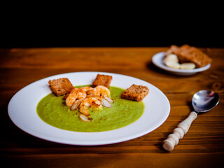 A plate of thick fresh green pea soup with prawns on a rustic wooden table with a garlic and croutons on the side.