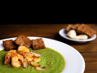 A plate of thick fresh green pea soup with prawns on a rustic wooden table with a garlic and croutons on the side. Close up.
