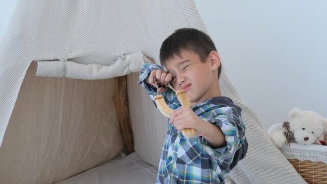 Little boy shooting with handmade slingshot in camera on wigwam background in light room