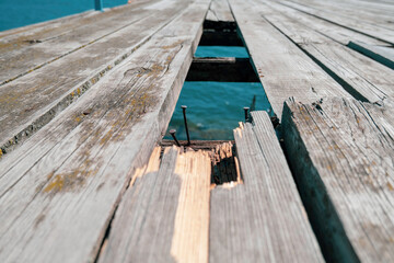 Old wooden bridge made of planks over the sea with a hole and rusty nails sticking out, through a hole in the floor you can see the blue sea