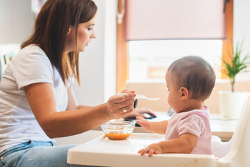 Obraz na płótnie Canvas Mother feed her little daughter with a spoon and search on internet food recipes