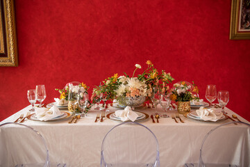 Elegant table setting with gold accents and red wall, clear chairs and flowers