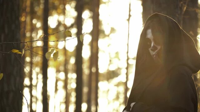 Witch in the hood and santa muerte makeup walking in autumn forest. The woman in black mantle is moving through trees in lights of sun. Theme of halloween and scary witchcraft the fall woods.