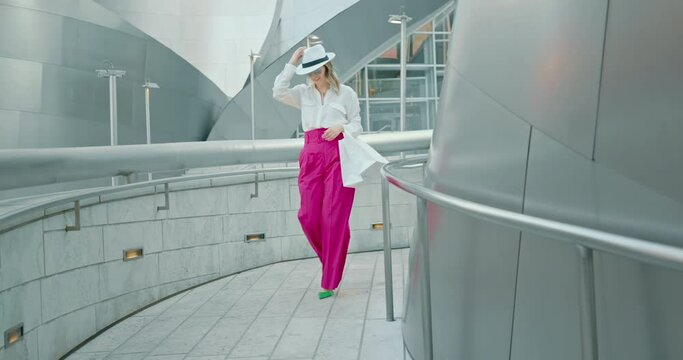 Pretty smiling blonde woman in stylish look confidently walks down the street with white shopping bags, touches her trendy white hat. Joy of life, lifestyle, fashion blogger, catwalk. 4K slow motion