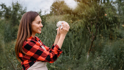 Young happy smiling woman farmer is holding a white baby rabbit in her hands and looking at it outdoors.