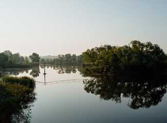 Dawn on the river. A fisherman is standing on the suspension bridge. Vegetation is reflected in calm water.