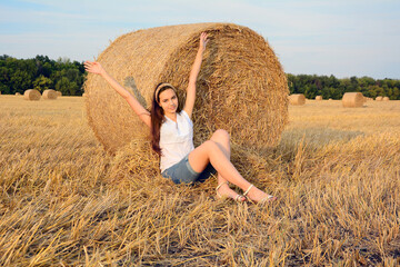 Naklejka na ściany i meble A young smiling girl sits next to a bale of straw with her hands up. Freedom and romance