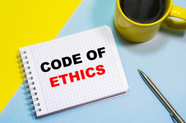 CODE OF ETHICS on a notepad on a blue-yellow background, a cup of coffee and a pen. Business concept