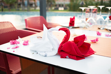 Red and white folded swan bird shape napkin in a restaurant on a white table at a resort near the pool. A flower petals and glasses on the table. Romantic dinner for newlyweds. Sunset.
