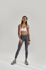 Full length shot of young sportive mixed race woman in sportswear looking at camera while posing isolated over grey background. Healthy lifestyle and sports concept