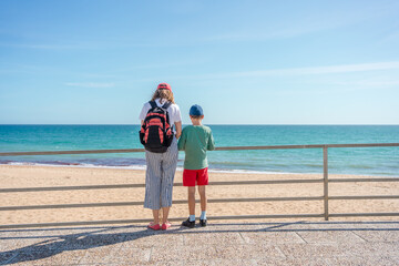 Mother with children standing by rail and relaxing looking at sea beach, sunshine outdoors background. Back view of recreation people dreaming romantic image.