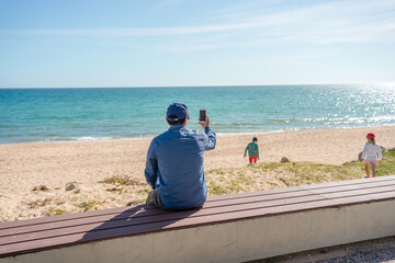 Back view of father taking sea beach pictures of kids by smart phone sunshine outdoors