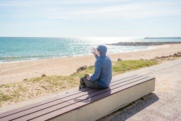 Back view of man drinking water sitting on sea view bench, sunny outdoors background
