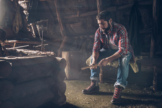 young bearded man sitting in wooden rustic alpine cabin and carving wood - Powered by Adobe