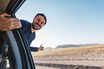 Young happy man getting out of the window of a car while driving in the middle of the desert on adventure travel