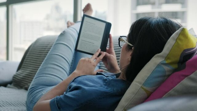 Happy female student reading ebook and lying on couch. Young woman relaxing with book. Caucasian girl and lifestyle, leisure and relaxation at home