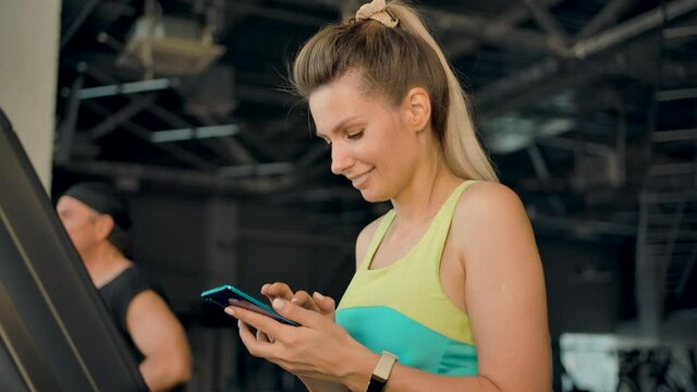 A pretty young blonde woman is walking on a treadmill in the gym and is typing a message on her mobile phone. She is smiling.