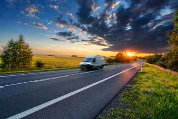 Motion blurred white fast delivery van driving on the asphalt road in rural landscape in the rays of the sunset with dark storm cloud