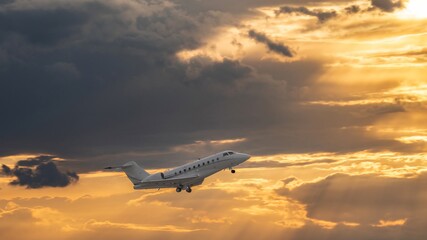 White small private turbofan-powered twin-engine business jet airplane flying in air after takeoff from airport. Dramatic orange grey cloudy sky at sunset after rain storm, dynamic weather conditions.