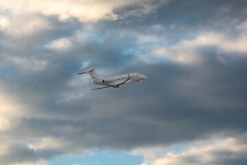 White small private turbofan-powered twin-engine business jet airplane flying in air after takeoff from airport. Dramatic orange grey cloudy sky at sunset after rain storm, dynamic weather conditions.