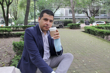 latin adult man with protection mask, drinking coffee in park, new normal covid-19
