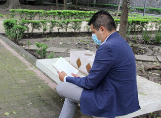 latin adult man with protection mask, reading book in park, new normal covid-19