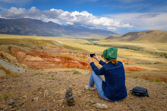 Young woman sitting on hill and taking photos on smartphone of a beautiful mountain landscape. Freedom, tourism, technology, adventure, discovery, travel for ordinary people. Image with copy space.