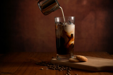 Glass of ice coffee while adding milk with biscuit and coffee beans on wooden table