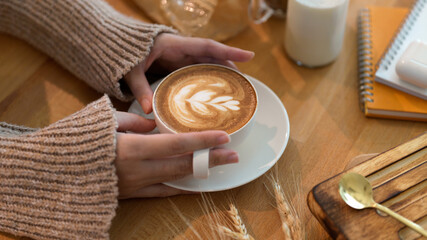 Cropped shot of female holding latte art coffee on wooden table with stationeries