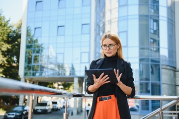 Successful female banker using tablet and wireless earphones outdoors near his office background lights, portrait young woman professional manager working on touch pad in evening city