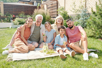 Portrait of modern multi-generation family having picnic on lawn in backyard on sunny summer day