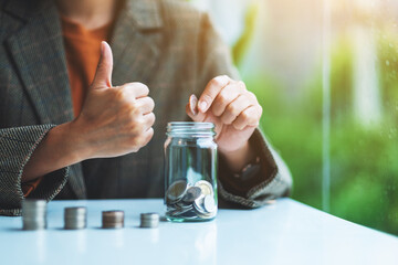 Closeup image of a businesswoman showing thumbs up hand sign while putting coins in a glass jar for saving money concept
