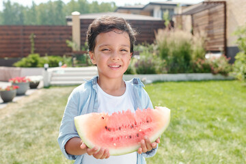 Waist up portrait shot of cheerful little boy standing in backyard holding piece of watermelon smiling at camera