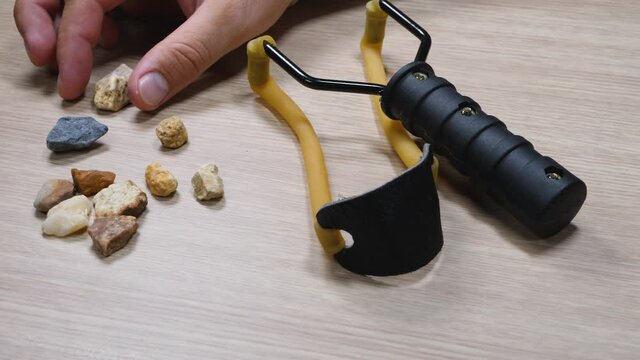 Man hand take stone with slingshot small hand-powered projectile weapon on wooden background. Classic form Y-shaped frame held in off hand with two rubber strips. Pocket that holds projectile
