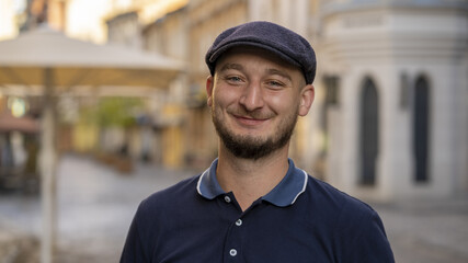 Street portrait of a smiling young man 30 -35 years old in a cap with a beard, looking directly into the camera on a neutral background of the old city.