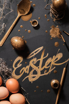Word ""Easter"" writtne with gold with painting set and eggs on black background..