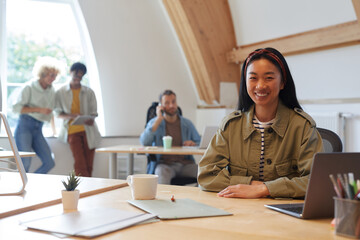 Portrait of Asian young woman smiling at camera while working at her workplace with other people in the background