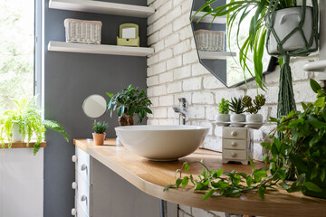 Stylish bathroom with white brick wall, mirror, green plants, washbasin and wooden furniture. Bright interior of spa in houseplant. 